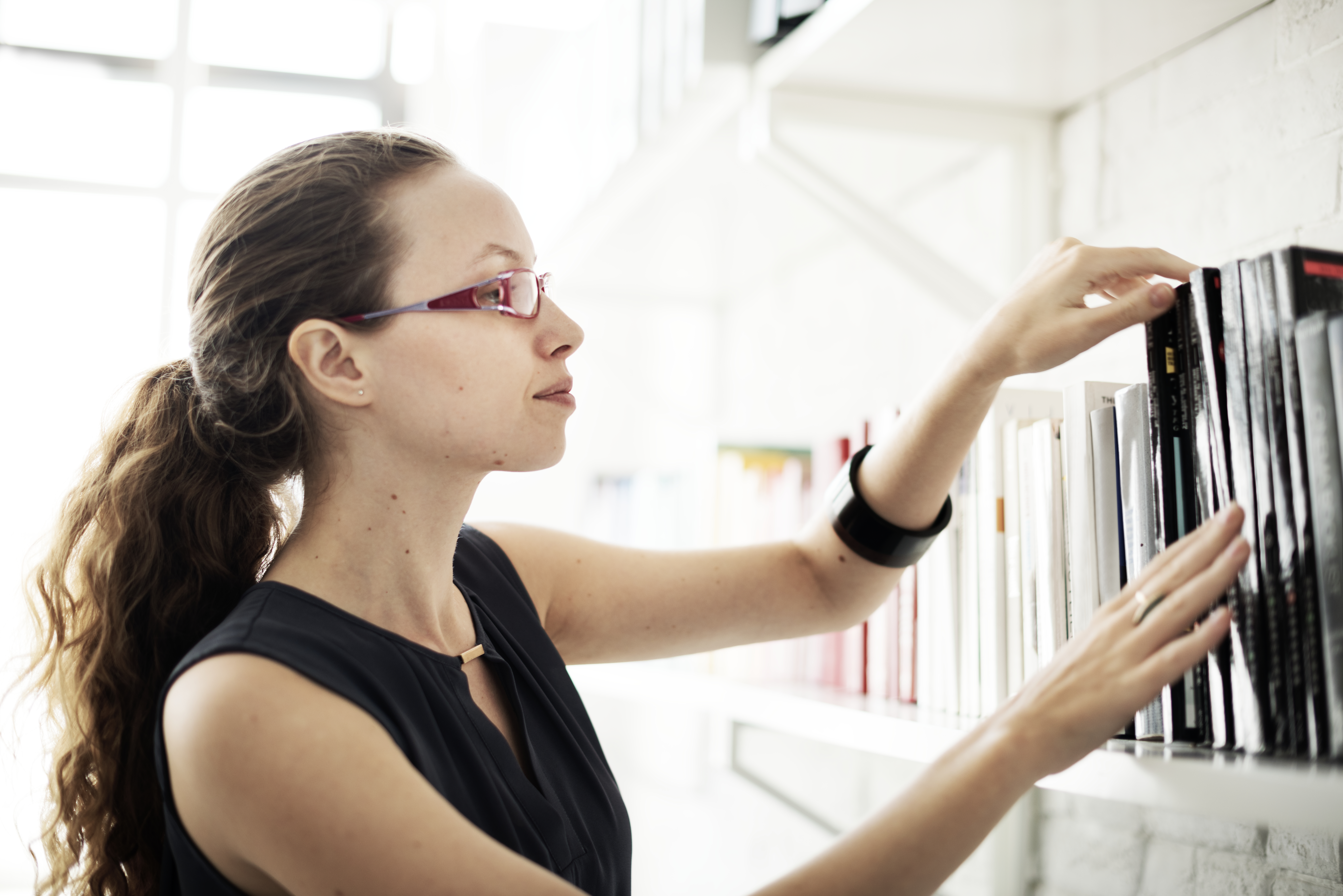 A woman in a black dress and glasses reaches out to printed books on shelf.