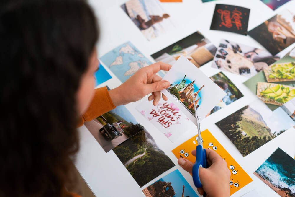 A woman sits at a table covered in photos and cuts out an image for a zine with scissors. 